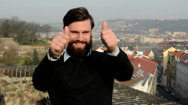 Młody przystojny mężczyzna z pełną brodą (hipster) pokazuje kciuki na temat porozumienia - miasto w tle — Wideo stockowe