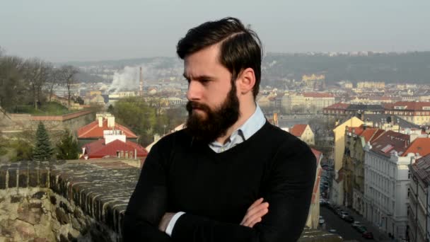 Young handsome man with full-beard (hipster) looks around and thinking (serious face) - city in background — Stock Video