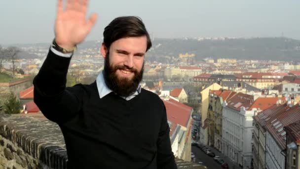 Jonge knappe man met volle baard (hipster) golven met de hand voor begroeting - stad op de achtergrond — Stockvideo