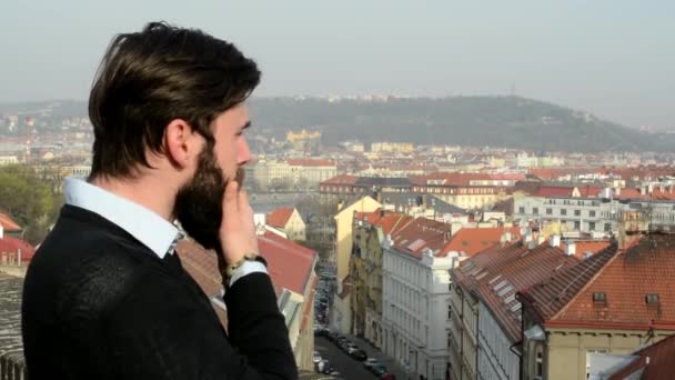 Joven hombre guapo con barba llena (hipster) mira a la ciudad y medita - ciudad en el fondo — Vídeo de stock