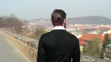 Genç, sakallı yakışıklı bir adam parkta yürüyor ve şehre bakıyor. Arka planda bir şehir var.