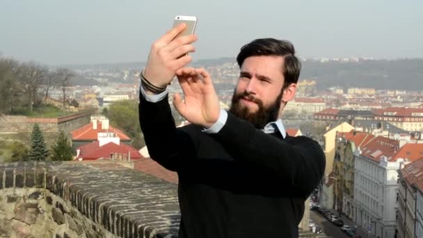 Νεαρός όμορφος άνδρας με πλήρη γενειάδα (hipster) παίρνει φωτογραφία (selfie) - πόλη στο παρασκήνιο — Αρχείο Βίντεο