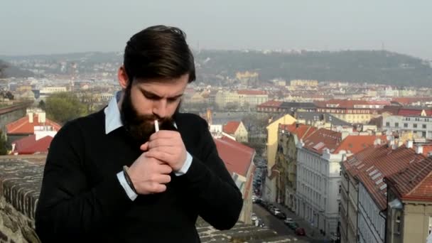 Ung stilig man med helskägg (hipster) rökning cigarett - stad i bakgrunden — Stockvideo