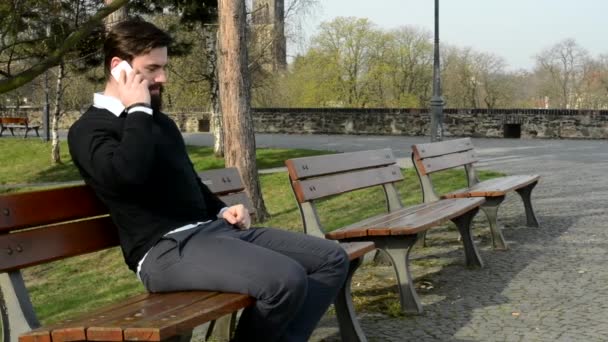 Jovem bonito homem com barba cheia (hipster) senta-se no banco e telefone - parque (natureza ) — Vídeo de Stock