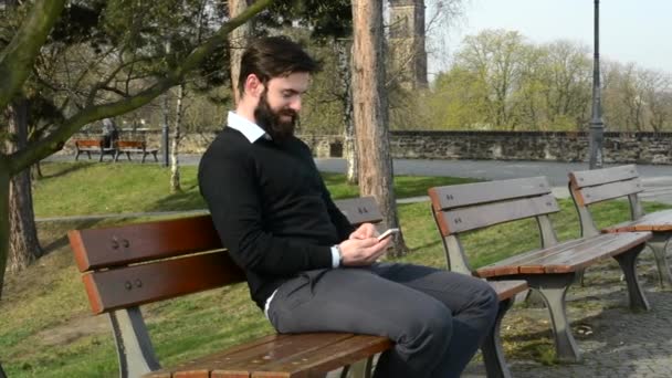 Fiatal jóképű férfi teljes szakállú (hipster) ül a padon, és dolgozik a mobiltelefon - park (természet) — Stock videók
