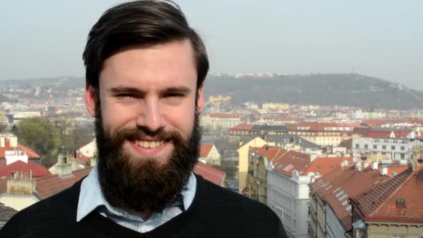 Young handsome man with full-beard (hipster) smiles to camera - city in background - closeup — Stock Video