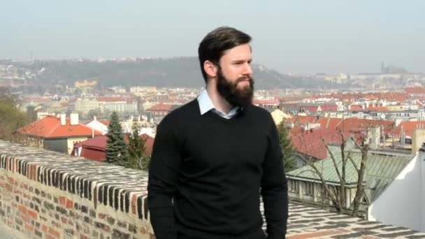 Jovem homem bonito com barba cheia (hipster) andando no parque e olha para a cidade - cidade no fundo — Vídeo de Stock