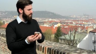 Genç ve sakallı bir adam cep telefonuyla yürüyor ve çalışıyor. Arka planda şehir (binalar - çatı -)