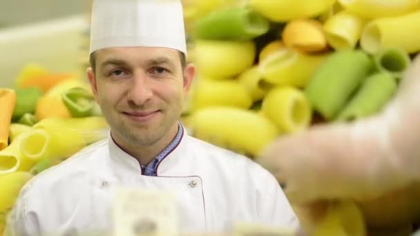 Chef sonríe a la cámara - pasta colorida seca en contenedor - primer plano - trabajador (manos) controla la calidad — Vídeos de Stock