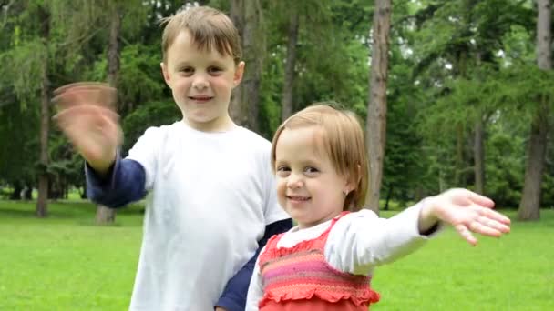 Niños (niño y niña (hermanos)) saludan con las manos y sonríen a la cámara - parque — Vídeo de stock