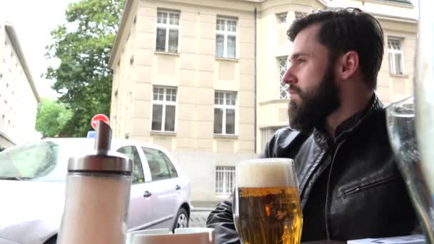 Joven hombre hipster guapo sentado en el restaurante (asientos al aire libre) y mira a su alrededor - calle - cerveza — Vídeos de Stock