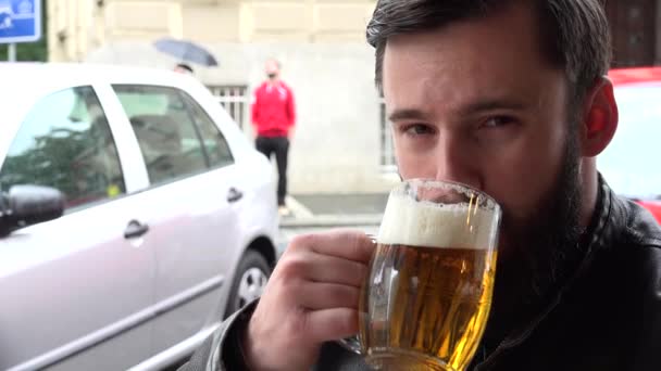 Joven guapo hipster hombre bebe cerveza en el restaurante y sonríe a la cámara - fuera de los asientos - calle con la gente y el coche aparcado — Vídeos de Stock