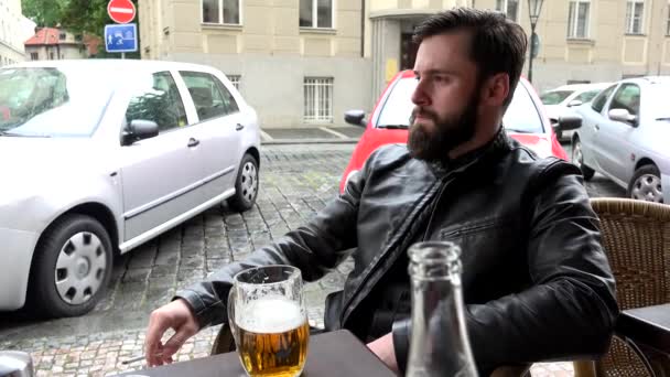 Junger gutaussehender Hipster-Mann sitzt im Restaurant (Sitzgelegenheiten im Freien) und raucht - Straße (geparkte Autos) - Bier — Stockvideo