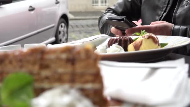 Jeune homme hipster beau travaille (dactylographier) sur smartphone - restaurant : sièges en plein air - rue avec voitures garées — Video