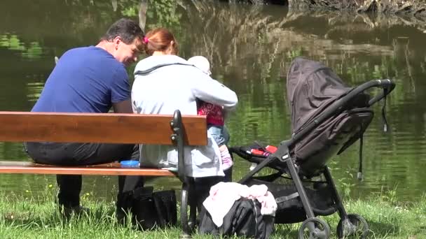 PRAGUE, REPÚBLICA CHECA - MAIO 31, 2015: família relaxar perto da lagoa - mather e pai com bebê sentar no banco - carrinho de bebê - close-up — Vídeo de Stock