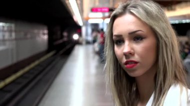 Genç ve çekici sarışın kadın metroyu bekliyor ve etrafına bakıyor. Arka planda diğer insanlar var.