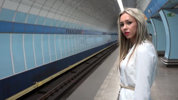 Joven mujer rubia atractiva espera el metro y mira a su alrededor - estación de metro - otras personas en el fondo — Vídeo de stock