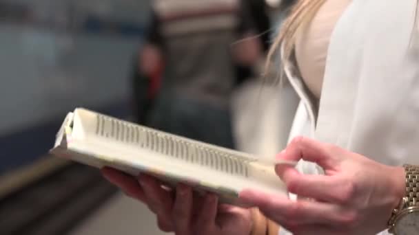 Joven mujer rubia atractiva lee un libro en el metro - estación de metro - otras personas en el fondo - manos de cerca — Vídeos de Stock