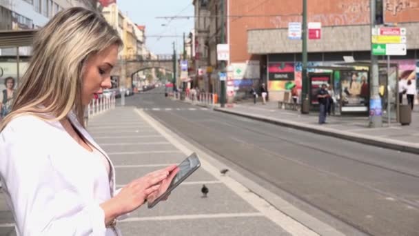 若い魅力的なブロンドの女性はタブレット上で動作します-街の人々と都市の通り — ストック動画