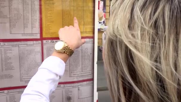 Молодая привлекательная блондинка смотрит на расписание (трамвай) и находит свою транзитную линию - выстрел сзади — стоковое видео