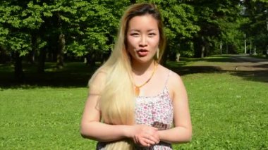 Genç, çekici, mutlu Asyalı kadın kameraya konuşur ve parkta el kol hareketi yapar.