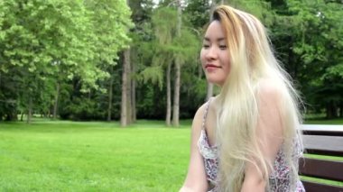 Genç Asyalı çekici mutlu kadın parkta bankta oturur - parkın etrafına bakın- yakın plan