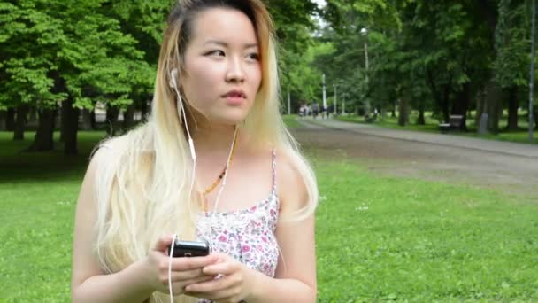 Молодая привлекательная счастливая азиатка слушает музыку на смартфоне в парке - наушники — стоковое видео