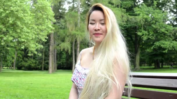 Junge asiatische attraktive glückliche Frau sitzt auf der Bank im Park - lächelt in die Kamera - Nahaufnahme — Stockvideo
