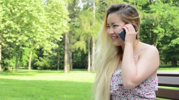 Jeune asiatique attrayant heureux femme s'assied sur le banc dans le parc téléphones avec quelqu'un gros plan — Video