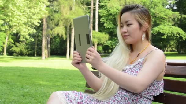 Jovem asiático atraente sorrisos mulher senta-se no banco no parque - tablet vídeo call - close-up — Vídeo de Stock