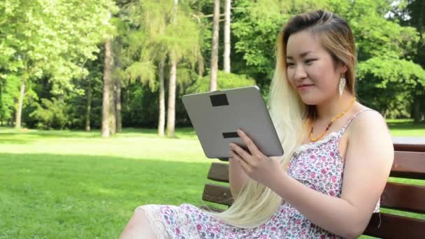 Молода азіатська приваблива щаслива жінка сидить на лавці в парку робота з планшетом крупним планом — стокове відео