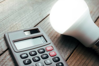 Hesap makinesi ve ahşap bir tahtanın üzerinde yanan bir ampul - Sağlayıcı Değişikliği Konseptiyle Enerji tasarrufu