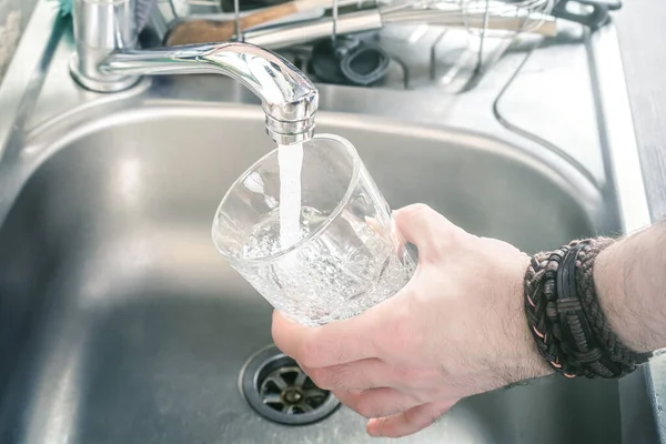 Mann hält Trinkglas über Küchenspüle und füllt es mit Wasser aus Wasserhahn — Stockfoto