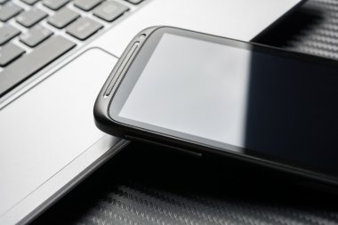Blank Business Smartphone With Reflection Leaning On Notebook Keyboard, All Above A Carbon Background