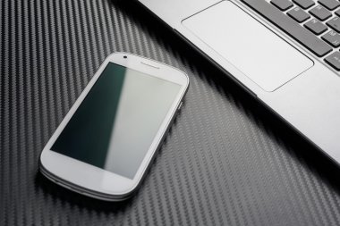 Blank Business Smartphone With Green Reflection Lying In Front Of A Notebook Touchpad, All Above A Carbon Background