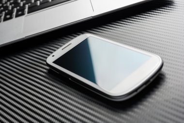 Blank Business Smartphone With Blue Reflection Lying In Front Of A Notebook Touchpad, All Above A Carbon Layer