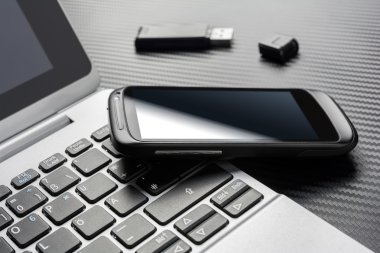 Blank Black Smartphone With Reflection Leaning On A Business Notebook Keyboard Next To An Open USB Storage Flash Drive, All Above A Carbon Layer