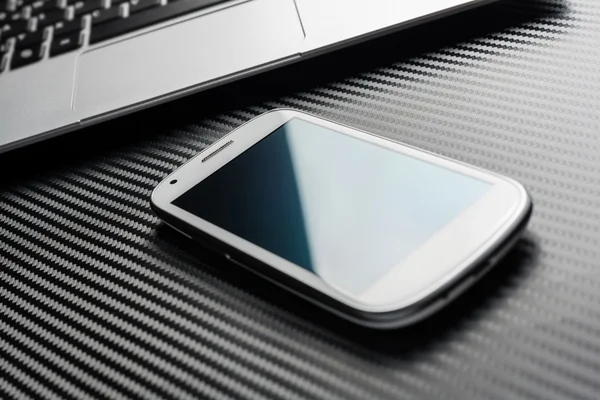 Blank Business Smartphone With Blue Reflection Lying In Front Of A Notebook Touchpad, All Above A Carbon Layer