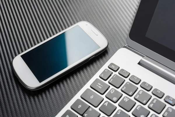 Geschäftsarbeit mit weißen, leeren Smartphones mit blauem Reflex, die auf einer Notebook-Tastatur liegen, alles über einer Carbonschicht — Stockfoto
