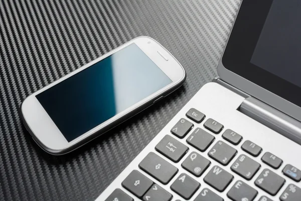 Business Work With White Blank Smartphone With Blue Reflection Lying Left To A Notebook Keyboard, All Above A Carbon Layer — Zdjęcie stockowe