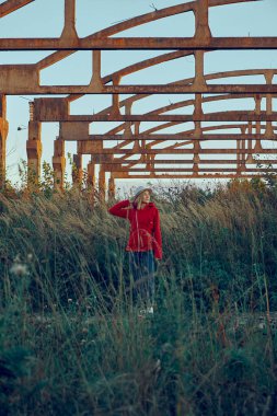 Kız açık havada duruyor, arka planda ise yapının bir iskeleti. Kırmızı ceketli ve şapkalı kız yan tarafa bakıyor. Dışarı çık. Yüksek kulakları olan düz bir arazi. Arkaplan