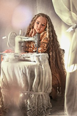 Retro giysili ve kıvırcık saçlı bebek hüzünle oturur ve yana bakar. Çocuğun önünde bir retro dikiş makinesi var. Dikiş işi kavramı, kendi ellerinle. Seçici odaklanma. retro