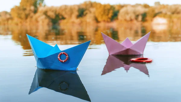 Deux bateaux en papier flottent sur la rivière, bleu et rose. Sur le bateau bleu pend une corde de sauvetage, et un autre flotte à côté de la rose Photo De Stock