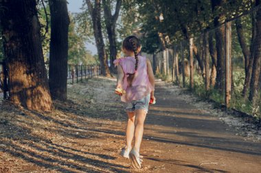 Bir kız bebek sokakta koşuyor, güneşli bir yaz günü, yol kenarları tüylerle kaplı, açık havada bir yürüyüş.