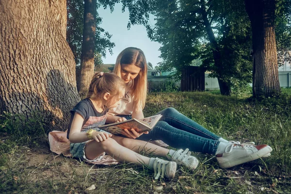 Mother and daughter read a book by the tree, on a sunny summer day. Outdoor recreation park.