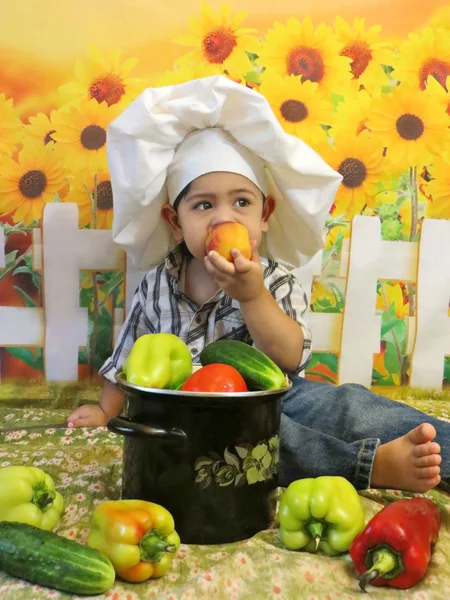 Menino sentado em um chapéu de chef com legumes come um pêssego em um fundo de girassóis — Fotografia de Stock