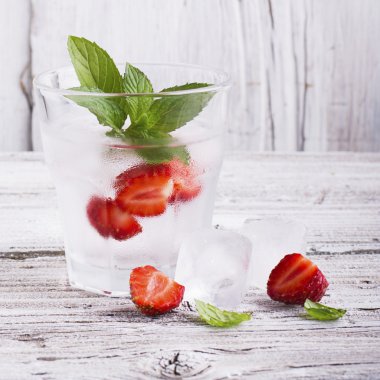 Basit bir beyaz ahşap arka plan içinde dilimleri sulu olgun çilek, nane yaprakları ve küpler ile ferahlatıcı buz su Cam. Konsept yaz tatili gevşemesi