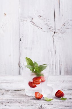 Basit bir beyaz ahşap arka plan içinde dilimleri sulu olgun çilek, nane yaprakları ve küpler ile ferahlatıcı buz su Cam. Konsept yaz tatili gevşemesi