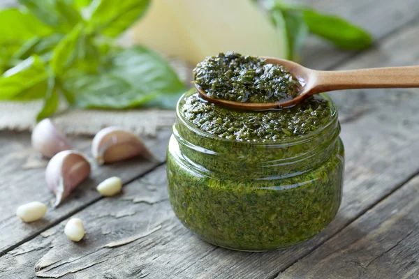 Gourmet traditional Italian pesto in glass jar with wooden spoon. Basil, nuts, olive oil and parmesan recipe. Vintage table background. — Stok fotoğraf