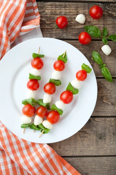 Seasonal traditional Italian caprese salad skewers with tomatoes basil and mozzarella cheese on white dish, rustic table background. — ストック写真